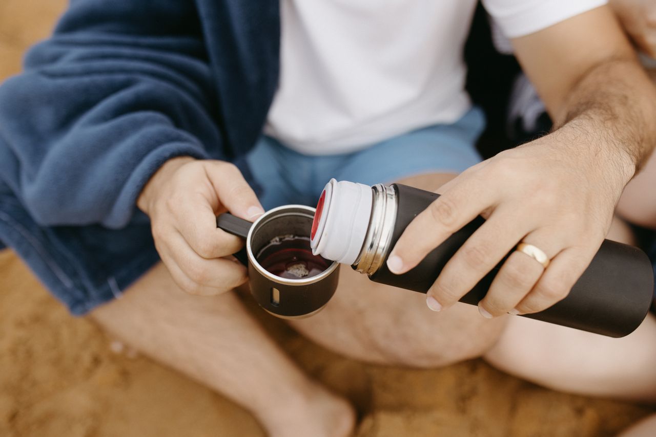 our-story A man in a robe pours a drink from a thermos into a mug on a sandy beach.