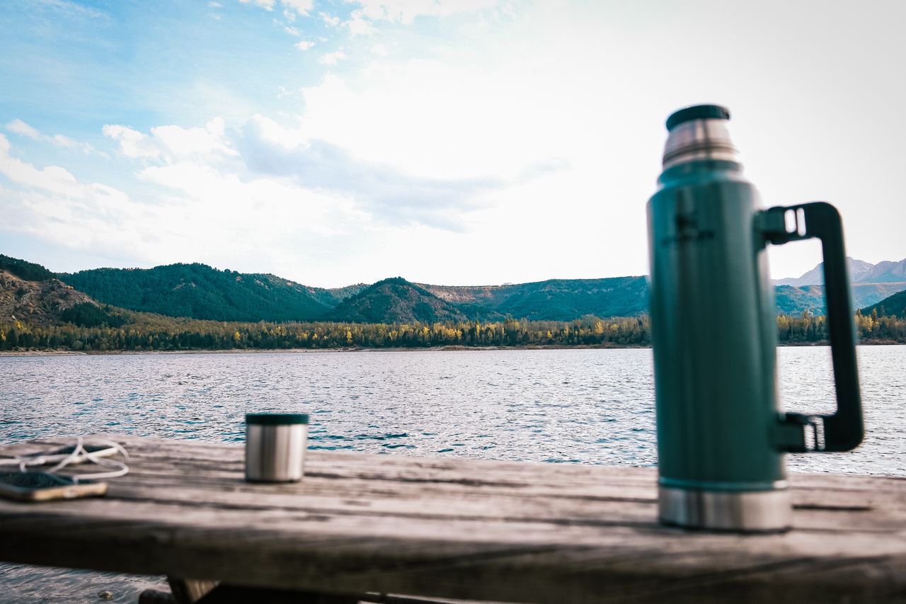 about-us A serene outdoor scene featuring a thermos and cup with a lake and mountains in the background.